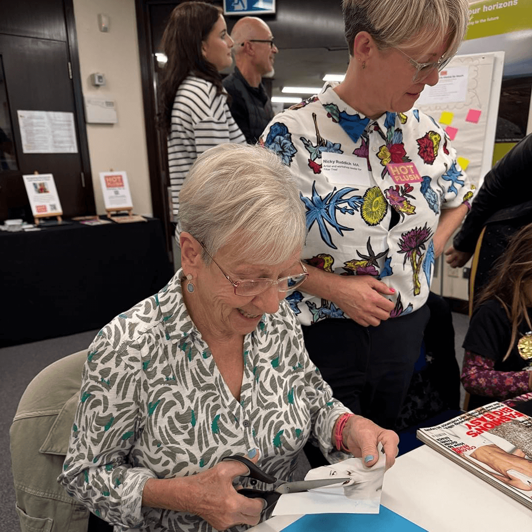A woman is seated at a table. She is cutting out an image from a magazine for a collage. A woman is seated at a table. She is cutting out an image from a magazine for a collage.