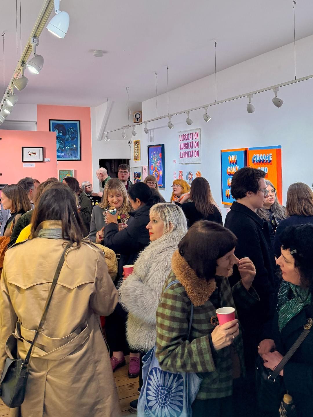 A room packed full of people mingle and enaged in talking. Some are holding pink cups. There are various artworks hung on the wall. A room packed full of people mingle and enaged in talking. Some are holding pink cups. There are various artworks hung on the wall.
