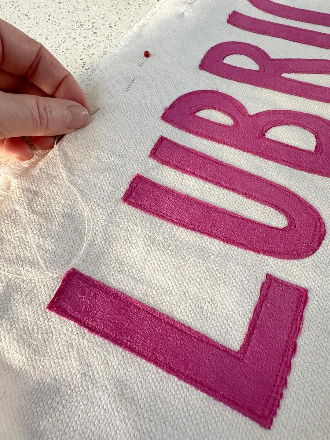 Close-up shot of cream linen fabric with bright pink cut out letters ‘L’ ‘U’ and ‘B’ sewn on. A hand is sewing the edge of the cream fabric. Close-up shot of cream linen fabric with bright pink cut out letters ‘L’ ‘U’ and ‘B’ sewn on. A hand is sewing the edge of the cream fabric.