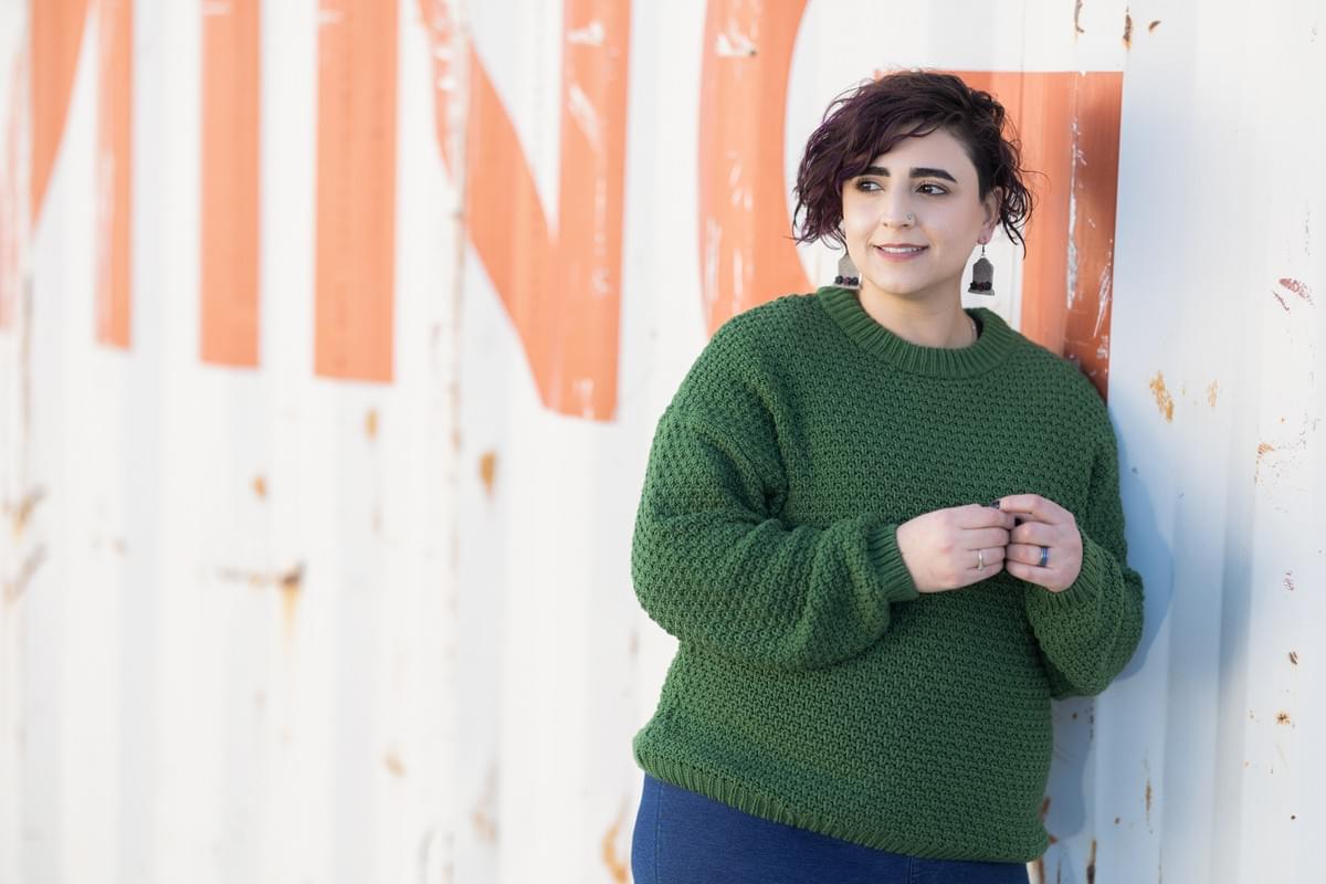 An image of Naseem, a light-skinned West Asian person with curly brown hair. They're leaning against a shipping container, hands fiddling, as they look off to the side. They're wearing tombstone earrings, a green sweater, and jeans, visible from the hips-up. An image of Naseem, a light-skinned West Asian person with curly brown hair. They're leaning against a shipping container, hands fiddling, as they look off to the side. They're wearing tombstone earrings, a green sweater, and jeans, visible from the hips-up.