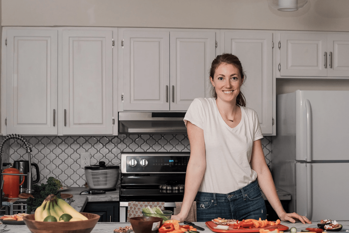 burnet woman standing in a white kitchen in a white tea shirt and blue lucky jeans, in front of a counter full of fresh fruit and veggies burnet woman standing in a white kitchen in a white tea shirt and blue lucky jeans, in front of a counter full of fresh fruit and veggies