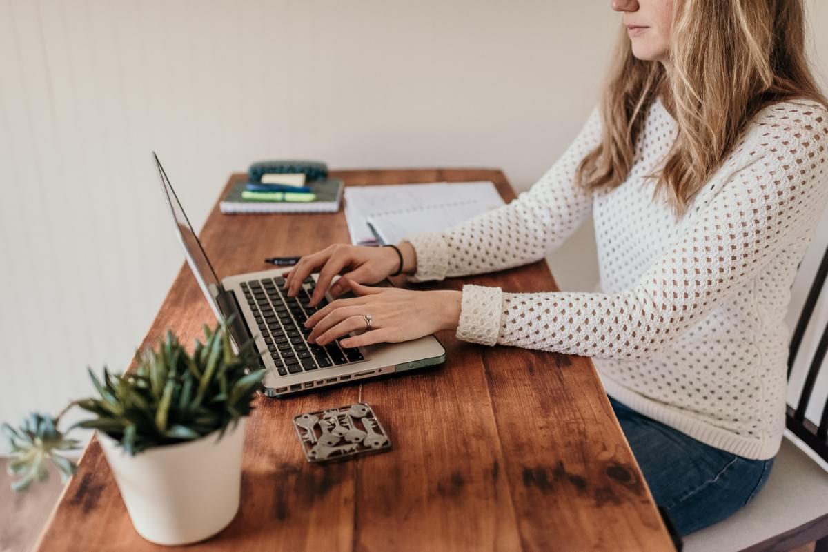 blond woman sitting at a wood desk in a white knit sweater typing on her Mac/ apple laptop blond woman sitting at a wood desk in a white knit sweater typing on her Mac/ apple laptop