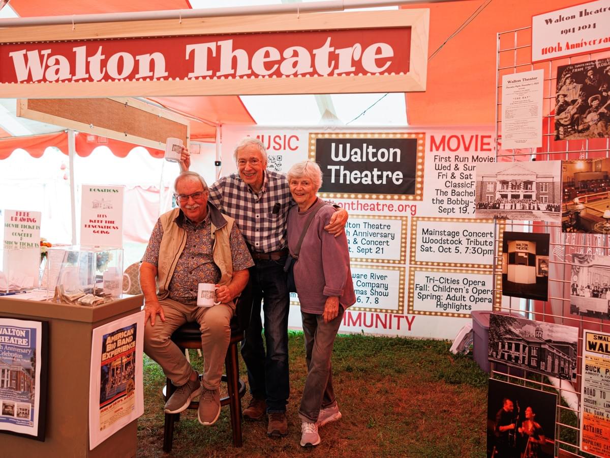 Photograph of three older adults posing inside a Walton Theatre promotional tent at an outdoor event, surrounded by posters and displays about upcoming movies, concerts, and theatre performances. The tent features a large red and white Walton Theatre banner, various black-and-white and color posters, and informational signs highlighting community events and theatre history. Photograph of three older adults posing inside a Walton Theatre promotional tent at an outdoor event, surrounded by posters and displays about upcoming movies, concerts, and theatre performances. The tent features a large red and white Walton Theatre banner, various black-and-white and color posters, and informational signs highlighting community events and theatre history.