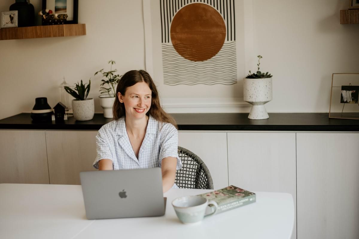 An image of Aleksa, a long haired woman in a blue dress, looking out of the frame and smiling at her desk. She is typing on a Macbook and has a tea cup and herbal book on the desk. There is a painting of a gold circle hung behind her. An image of Aleksa, a long haired woman in a blue dress, looking out of the frame and smiling at her desk. She is typing on a Macbook and has a tea cup and herbal book on the desk. There is a painting of a gold circle hung behind her.