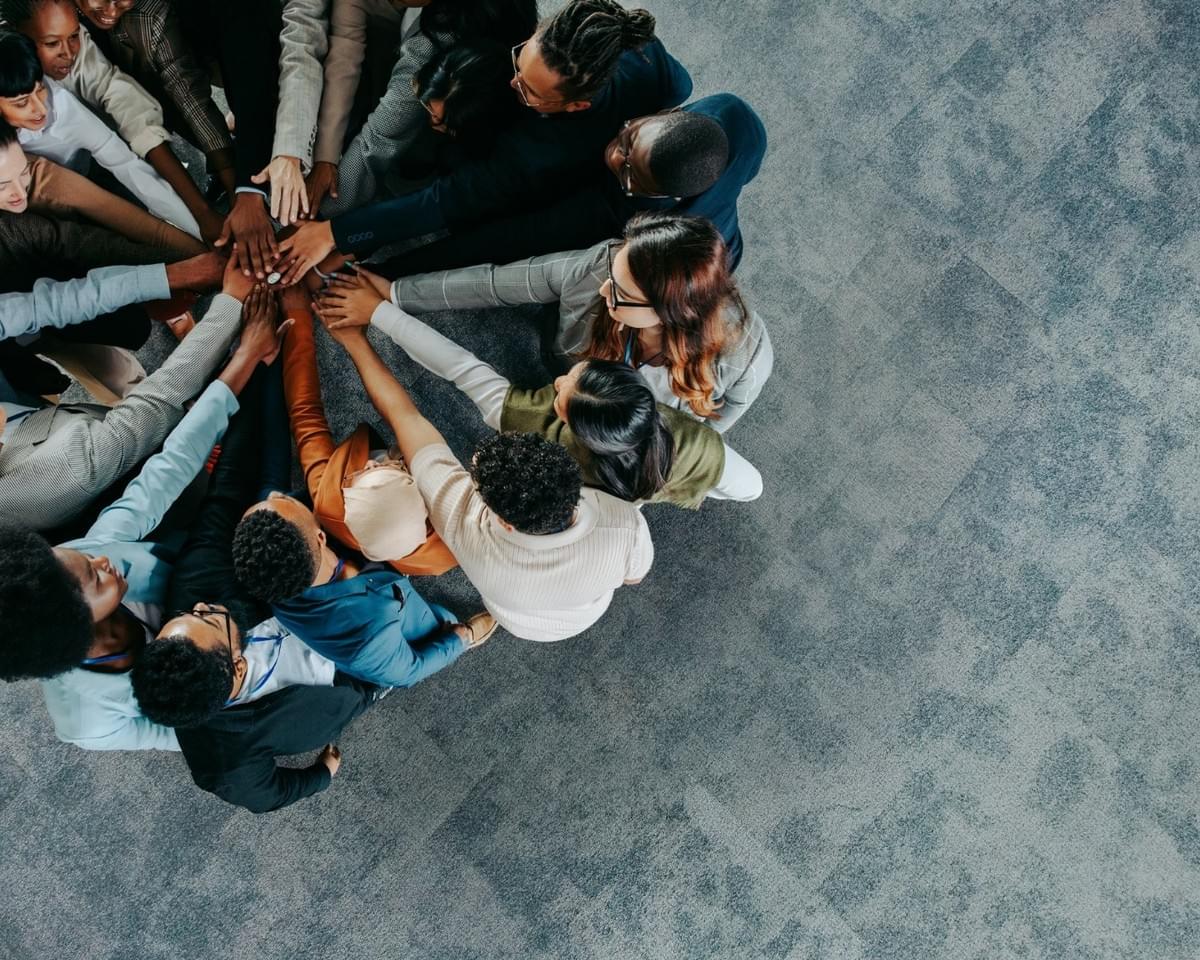 A diverse group of people stand in a circle with their hands stacked together in the center, viewed from above. The image emphasizes unity, teamwork, and collective effort against a grey carpeted background A diverse group of people stand in a circle with their hands stacked together in the center, viewed from above. The image emphasizes unity, teamwork, and collective effort against a grey carpeted background