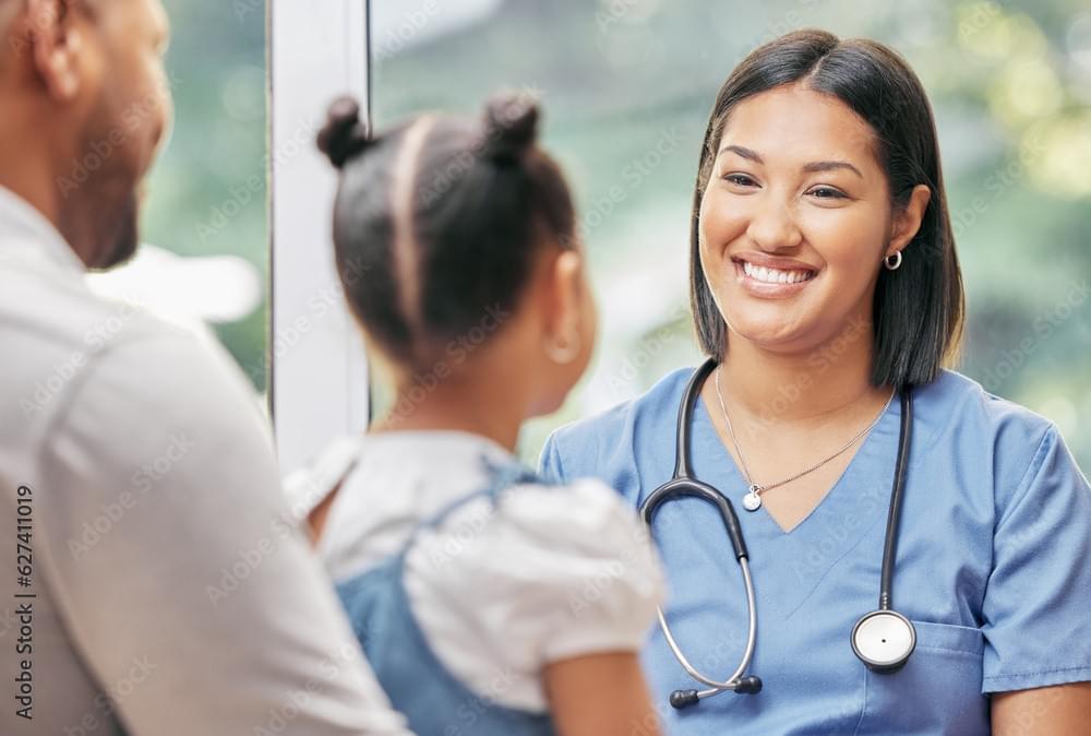 A healthcare professional in blue scrubs with a stethoscope around the neck speaks with a young girl who has her hair in two buns. The girl is accompanied by an adult, and they are in a bright room with large windows in the background. A healthcare professional in blue scrubs with a stethoscope around the neck speaks with a young girl who has her hair in two buns. The girl is accompanied by an adult, and they are in a bright room with large windows in the background.