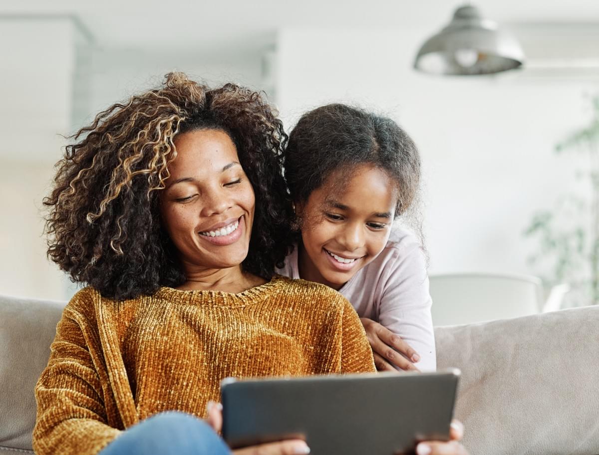 A mother sits on a couch wearing a mustard-colored sweater and holds a tablet, while her daughter leans over from behind in a light-colored shirt, both focused on the screen. A modern light fixture and greenery are visible in the background A mother sits on a couch wearing a mustard-colored sweater and holds a tablet, while her daughter leans over from behind in a light-colored shirt, both focused on the screen. A modern light fixture and greenery are visible in the background