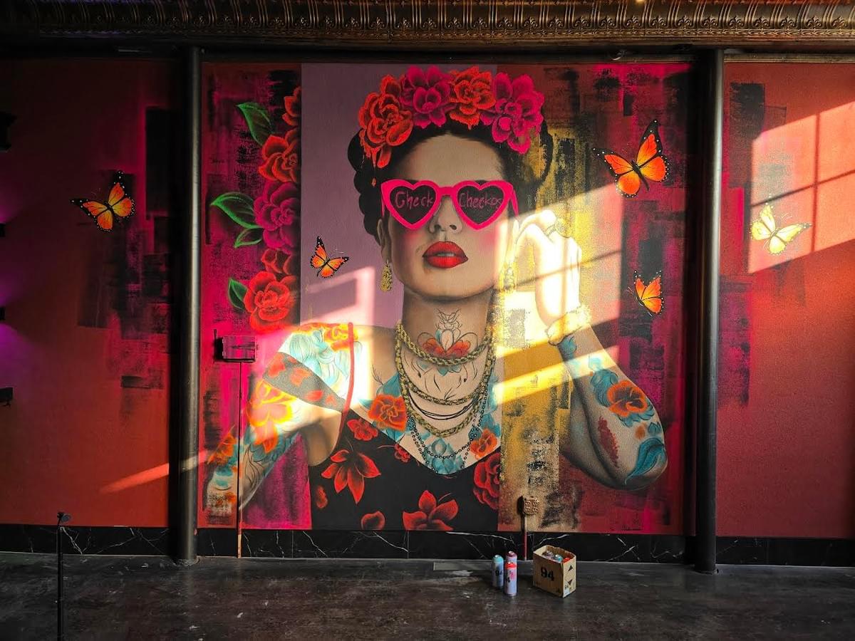 Large contemporary mural by Sergio Martinez “Checkos” featuring a stylized Frida Kahlo portrait with heart shaped sunglasses, floral tattoos, butterflies, and neon pink tones inside an interior space. Large contemporary mural by Sergio Martinez “Checkos” featuring a stylized Frida Kahlo portrait with heart shaped sunglasses, floral tattoos, butterflies, and neon pink tones inside an interior space.