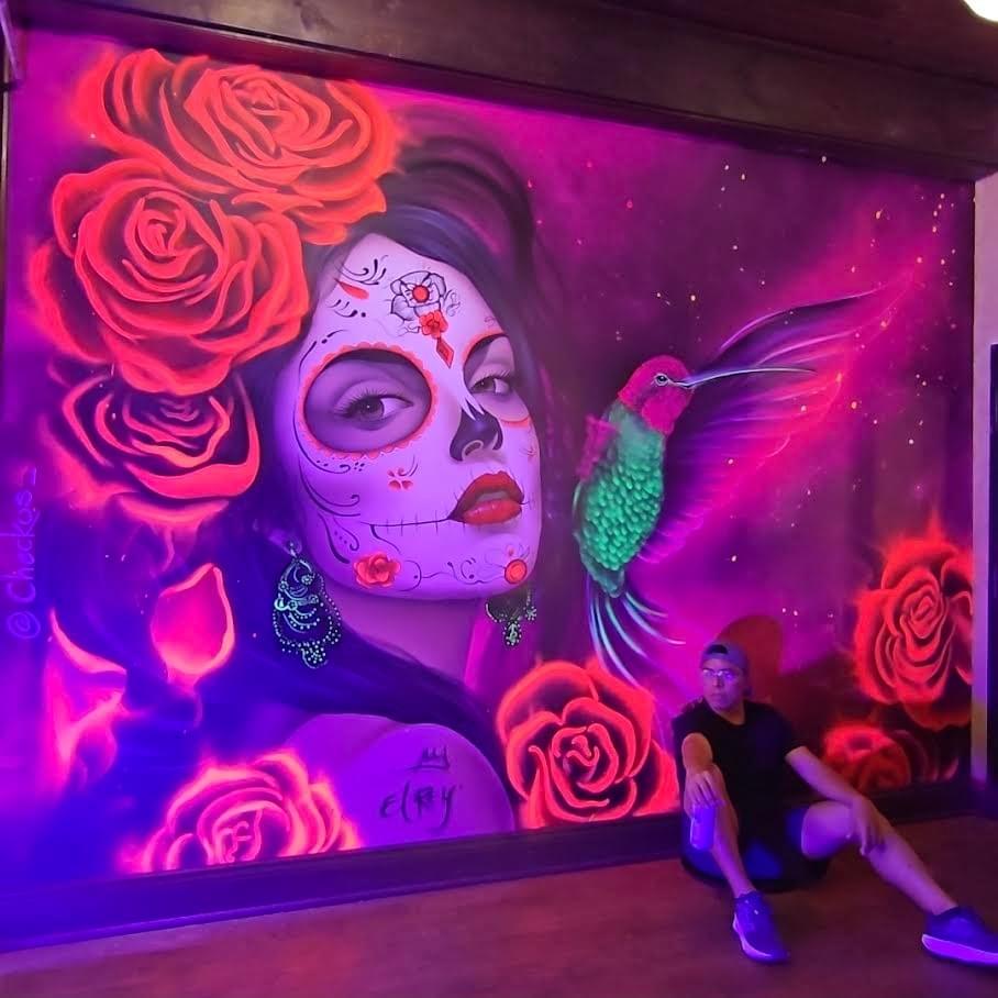 Vibrant neon mural by California artist Sergio Martinez “Checkos” featuring a Día de los Muertos inspired Catrina portrait surrounded by glowing roses and a hummingbird against a purple cosmic background. Vibrant neon mural by California artist Sergio Martinez “Checkos” featuring a Día de los Muertos inspired Catrina portrait surrounded by glowing roses and a hummingbird against a purple cosmic background.