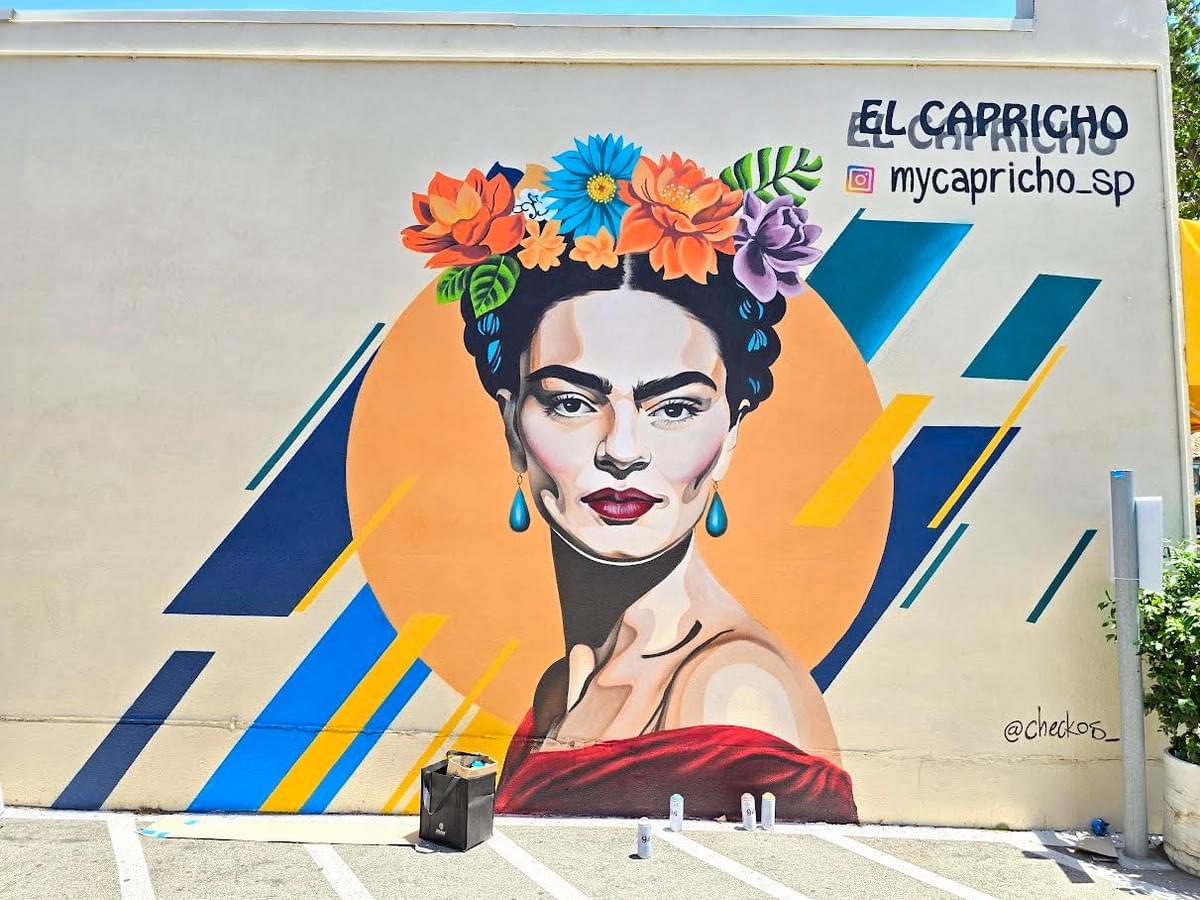 Colorful mural by California artist Sergio Martinez “Checkos” featuring a stylized portrait of Frida Kahlo with floral crown imagery and geometric background elements painted on an outdoor wall. Colorful mural by California artist Sergio Martinez “Checkos” featuring a stylized portrait of Frida Kahlo with floral crown imagery and geometric background elements painted on an outdoor wall.