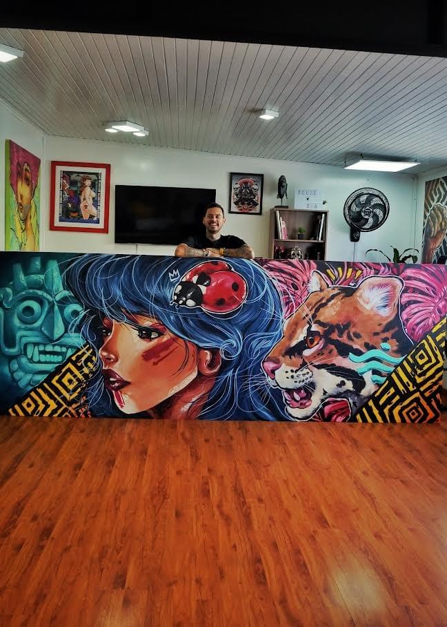 Daniel Acosta León posing behind a large original painting featuring a woman with blue hair, a jaguar, a ladybug, and symbolic Colombian inspired imagery rendered in bold, vibrant colors. Daniel Acosta León posing behind a large original painting featuring a woman with blue hair, a jaguar, a ladybug, and symbolic Colombian inspired imagery rendered in bold, vibrant colors.