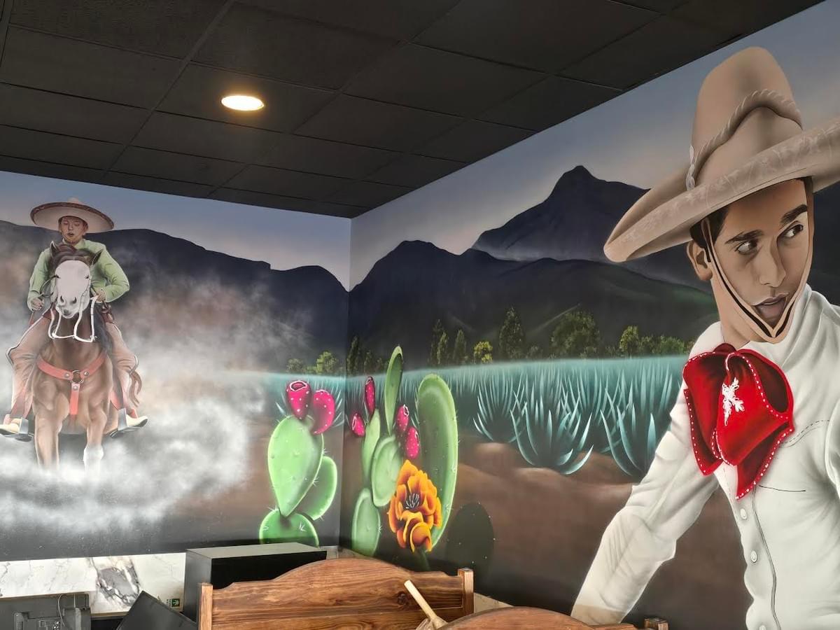 Large mural by California artist Sergio Martinez “Checkos” featuring charro inspired figures, agave fields, cactus plants, and mountainous landscapes painted across an interior restaurant wall celebrating Mexican culture and heritage. Large mural by California artist Sergio Martinez “Checkos” featuring charro inspired figures, agave fields, cactus plants, and mountainous landscapes painted across an interior restaurant wall celebrating Mexican culture and heritage.