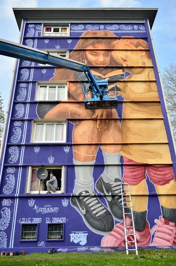 Large scale mural by Tlacolulokos painted on a purple apartment building, featuring two children reading together and blending themes of youth, identity, and urban Indigenous culture within a contemporary city setting Large scale mural by Tlacolulokos painted on a purple apartment building, featuring two children reading together and blending themes of youth, identity, and urban Indigenous culture within a contemporary city setting