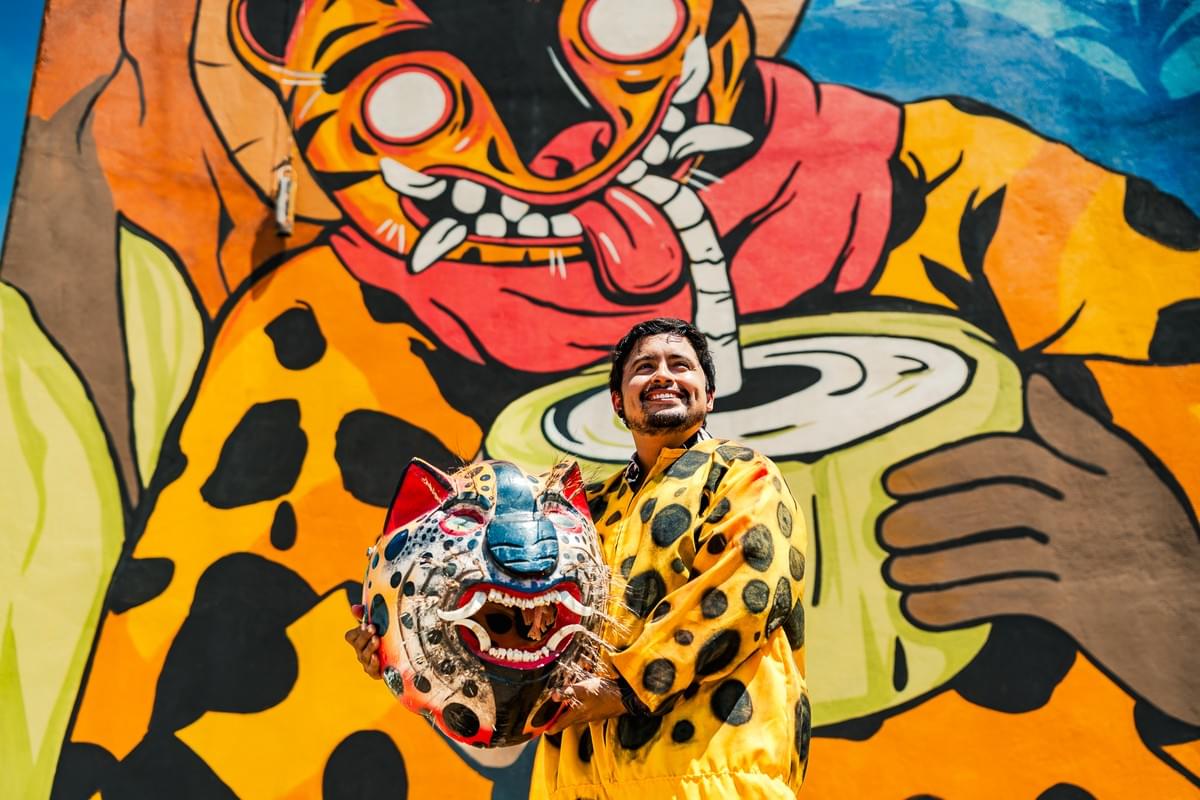 Mexican artist Miguel Castillo posing in front of a colorful jaguar inspired mural while holding a traditional feline mask connected to Mexican folklore and coastal culture. Mexican artist Miguel Castillo posing in front of a colorful jaguar inspired mural while holding a traditional feline mask connected to Mexican folklore and coastal culture.
