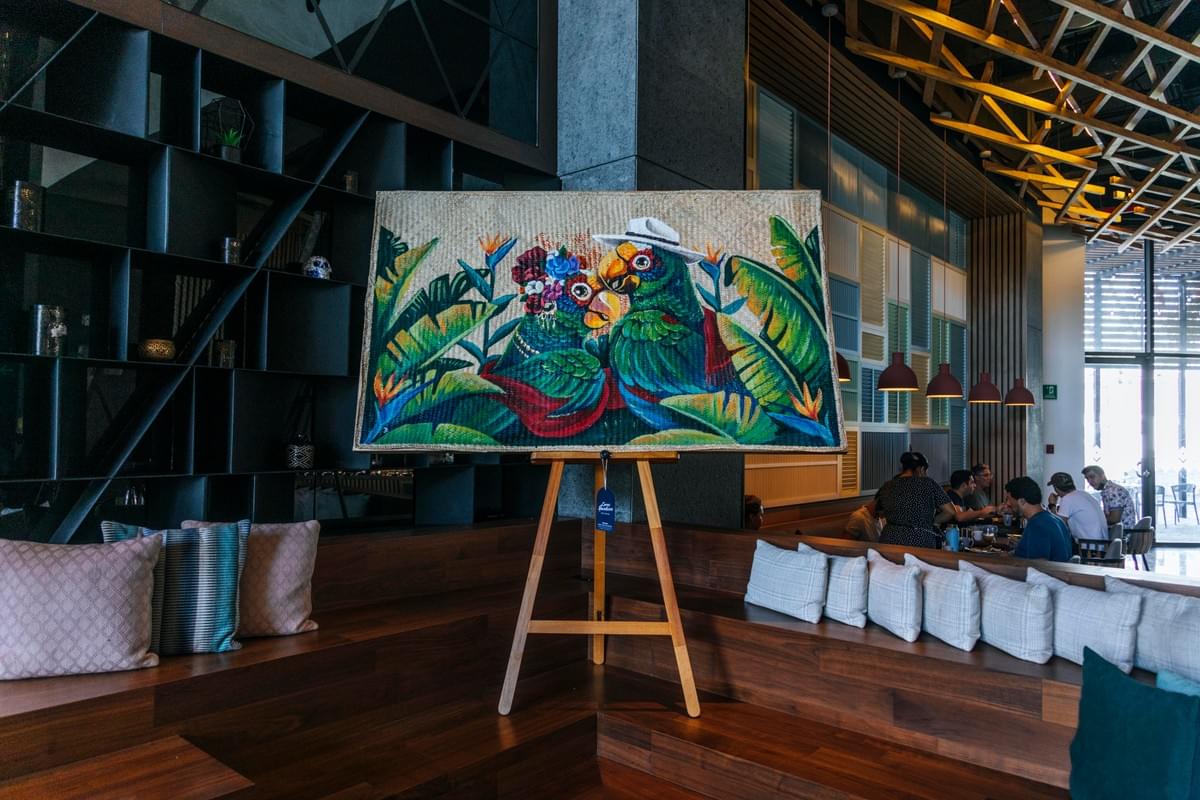 Colorful tropical painting by Miguel Castillo featuring two parrots surrounded by lush jungle leaves and coastal inspired floral patterns displayed inside a modern interior space. Colorful tropical painting by Miguel Castillo featuring two parrots surrounded by lush jungle leaves and coastal inspired floral patterns displayed inside a modern interior space.