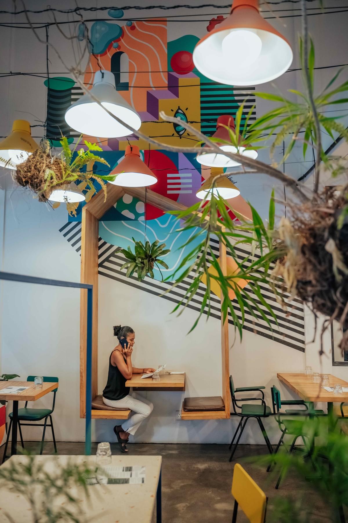 Espace de restauration intérieur de Nouvelle Usine à Floréal, aménagé avec une fresque colorée, des plantes et des luminaires suspendus. Espace de restauration intérieur de Nouvelle Usine à Floréal, aménagé avec une fresque colorée, des plantes et des luminaires suspendus.