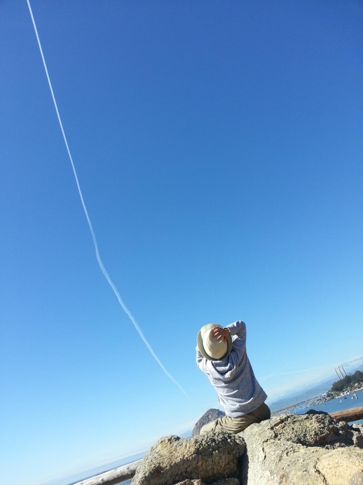 boy in a hat looking up at airplane contrail against a blue sky with Morro Bay, CA in the background. Photo credit: Laurie Ferguson boy in a hat looking up at airplane contrail against a blue sky with Morro Bay, CA in the background. Photo credit: Laurie Ferguson