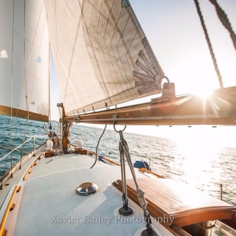 Photo description: a wooden sailboat on the ocean with the sun shining behind the sail, Xavier Bailey Photography Photo description: a wooden sailboat on the ocean with the sun shining behind the sail, Xavier Bailey Photography