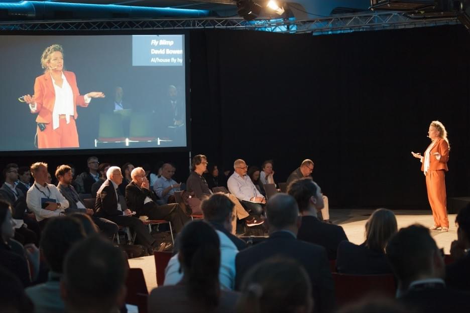 Dr. Selin stands at the right side of the image, on a stage. Facing her, on the left, is a screen that shows her in a zoomed in image from the front. Between Dr. Selin and the screen, an audience sits, facing her on the stage. Dr. Selin stands at the right side of the image, on a stage. Facing her, on the left, is a screen that shows her in a zoomed in image from the front. Between Dr. Selin and the screen, an audience sits, facing her on the stage.