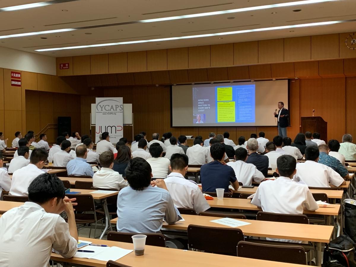 Photo taken from the back of a seminar hall, where many people seated at tables quietly listen to a speaker at the front of the room Photo taken from the back of a seminar hall, where many people seated at tables quietly listen to a speaker at the front of the room