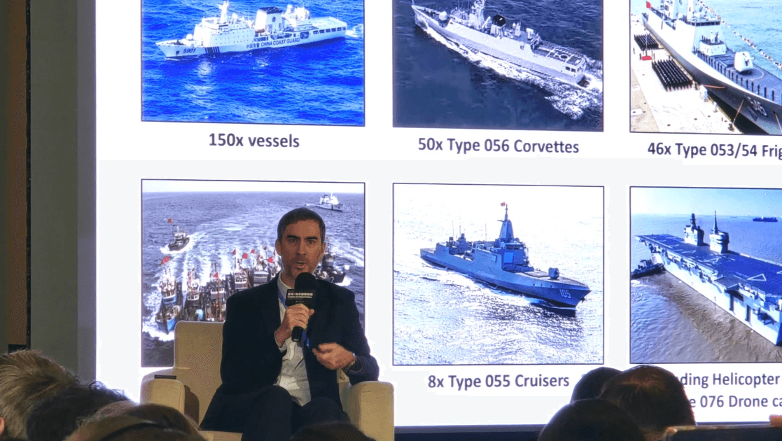 Benjamin Blandin holds a microphone while sitting in a plush panelist chair, speaking with focus. Behind him is a slide display photos of six types of Chinese coast guard, naval, and militia ships/boats, detailing how many of each China has. Benjamin Blandin holds a microphone while sitting in a plush panelist chair, speaking with focus. Behind him is a slide display photos of six types of Chinese coast guard, naval, and militia ships/boats, detailing how many of each China has.