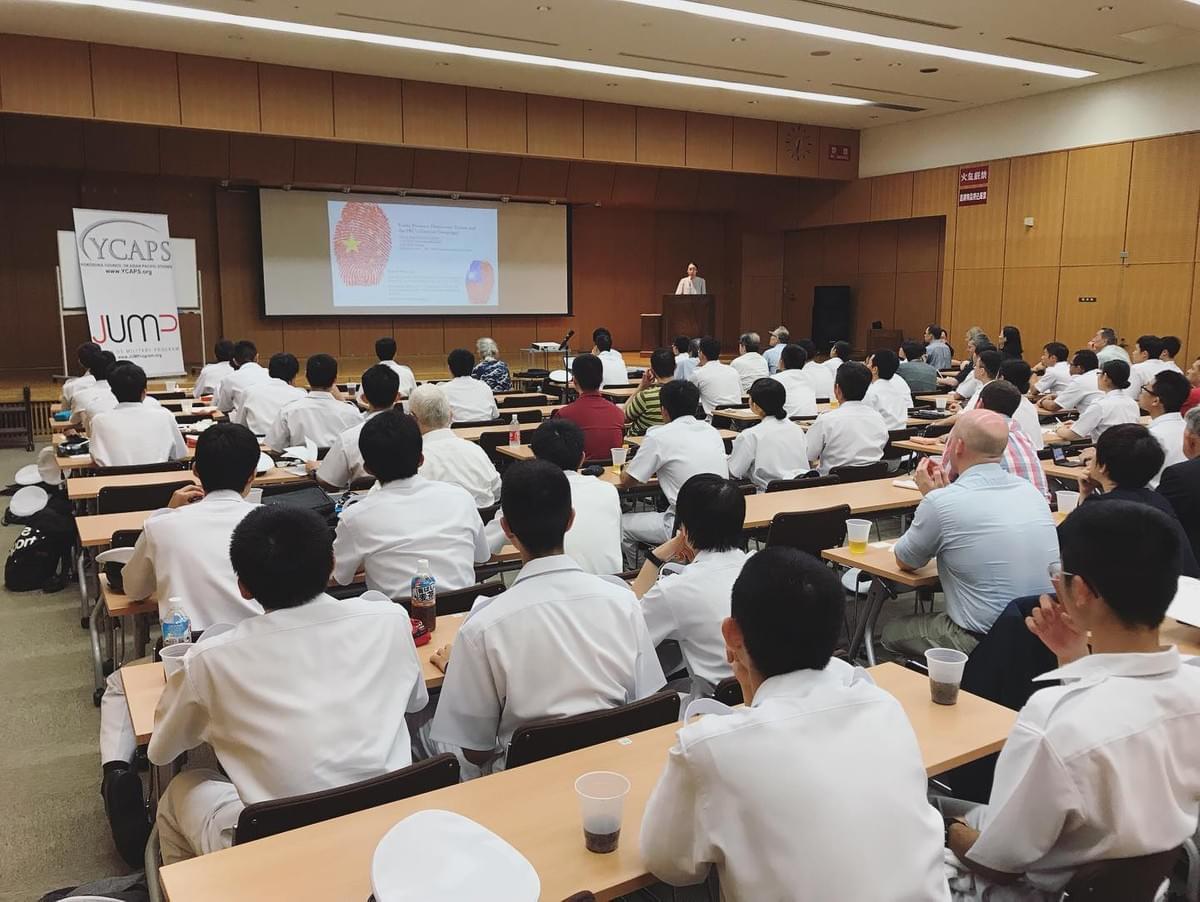 Photo taken from the back of a seminar hall where many attendees are quietly watching a presenter at the front of the room Photo taken from the back of a seminar hall where many attendees are quietly watching a presenter at the front of the room
