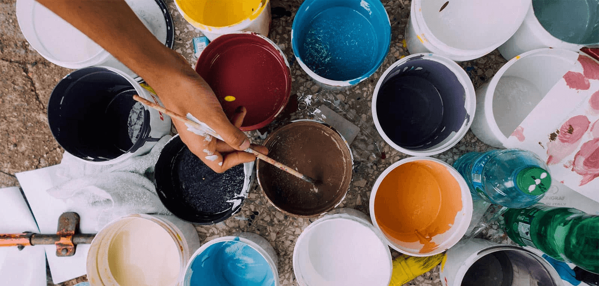 A brown-skinned person's hand holding a paintbrush and surrounded by buckets of paint. A brown-skinned person's hand holding a paintbrush and surrounded by buckets of paint.