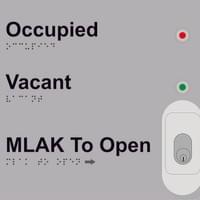 External door control panel, with braille and tactile text saying "Occupied", "Vacant" and "MLAK to Open". There is a keyhole for the MLAK. There is a red light next to "Occupied" and a green light next to "Vacant". There is a braille equivalent below all tactile text.