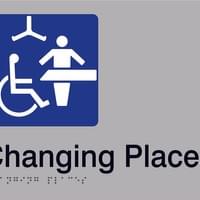The Changing Places Logo on a braille and tactile sign, with the words "Changing Places". The Changing Places logo is a pictogram of a wheelchair user, next to an adult-sized change table and a person standing next to it. The is an overhead ceiling hoist too. 