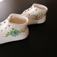The left hand side of two porcelain baby shoes showing the delicately painted flows and foliage. One has a blue forget-me-nota and one has a pink rose.