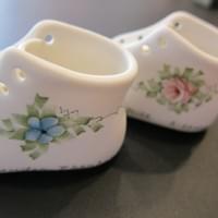 The left hand side of two porcelain baby shoes showing the delicately painted flows and foliage. One has a blue forget-me-nota and one has a pink rose.