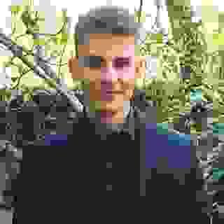 A young man in his 20s has a pleasant neutral expression, wearing a black suit and black shirt, standing in front of some ivy-covered trees and bushes. He has pale skin, dark eyes, and short blond hair. A young man in his 20s has a pleasant neutral expression, wearing a black suit and black shirt, standing in front of some ivy-covered trees and bushes. He has pale skin, dark eyes, and short blond hair.