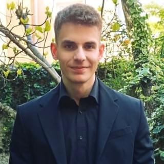 A young man in his 20s has a pleasant neutral expression, wearing a black suit and black shirt, standing in front of some ivy-covered trees and bushes. He has pale skin, dark eyes, and short blond hair.