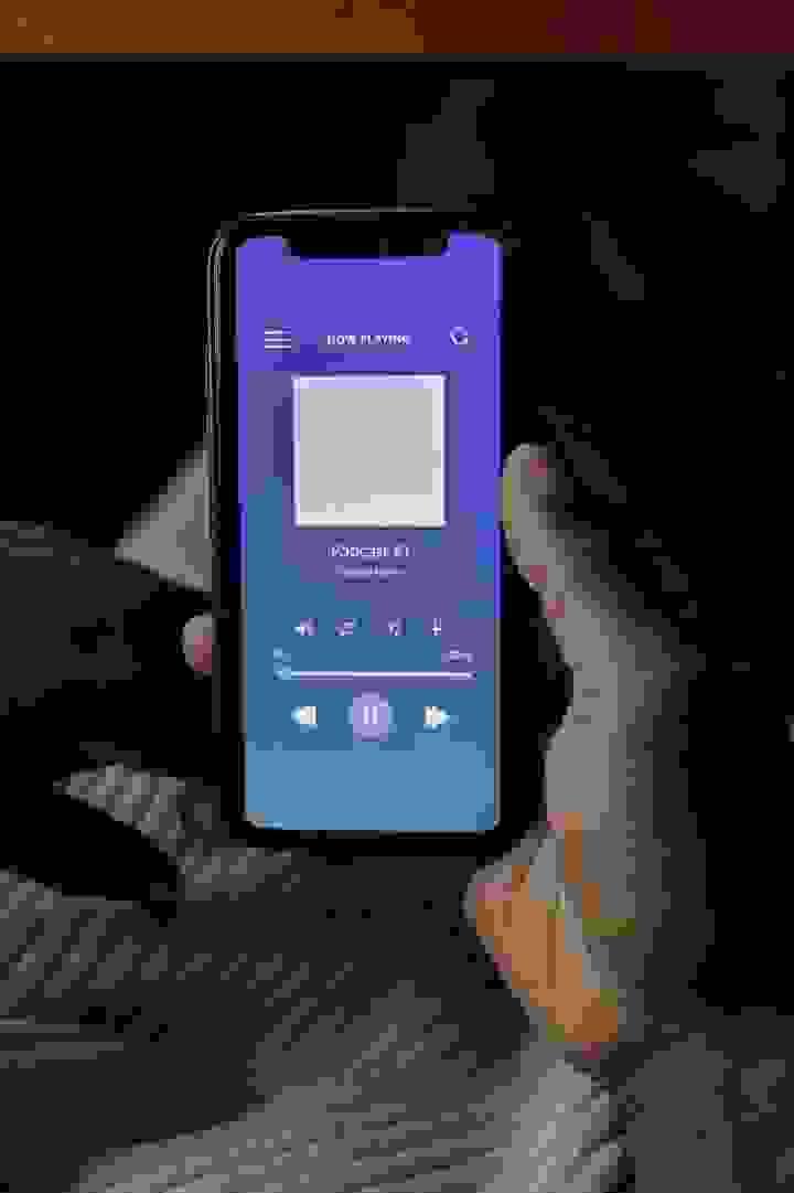 A person holds a smartphone displaying a podcast player interface. The screen shows “NOW PLAYING” at the top, followed by the title “Podcast #1” and the subtitle “Podcast name.” Standard media controls—play/pause, skip forward, and skip backward—are visible beneath the text, along with a playback progress bar. The app’s background features a smooth gradient of blue and purple hues, creating a calm and modern aesthetic. The image captures the experience of engaging with digital audio content, emphasizing themes of mobile accessibility, personal enrichment, and the integration of technology into everyday listening habits A person holds a smartphone displaying a podcast player interface. The screen shows “NOW PLAYING” at the top, followed by the title “Podcast #1” and the subtitle “Podcast name.” Standard media controls—play/pause, skip forward, and skip backward—are visible beneath the text, along with a playback progress bar. The app’s background features a smooth gradient of blue and purple hues, creating a calm and modern aesthetic. The image captures the experience of engaging with digital audio content, emphasizing themes of mobile accessibility, personal enrichment, and the integration of technology into everyday listening habits