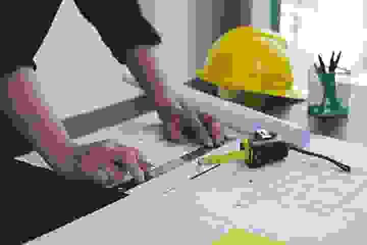 Two people are seated at a desk and are reviewing a set of building plans, the desk also has a laptop, level, and hardhat on it Two people are seated at a desk and are reviewing a set of building plans, the desk also has a laptop, level, and hardhat on it