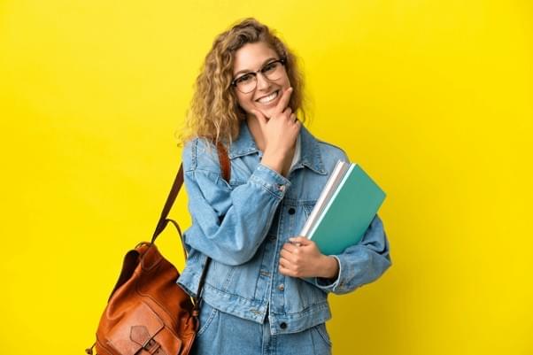 Young woman wearing glasses and a leather bag holding text books and smiling - representing online therapy with pricing that would be affordable for students. Young woman wearing glasses and a leather bag holding text books and smiling - representing online therapy with pricing that would be affordable for students.