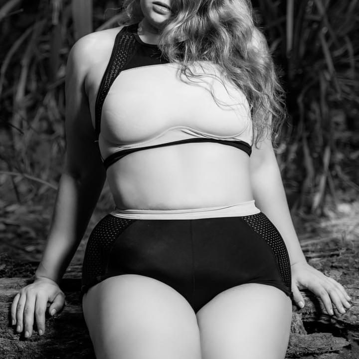 Black and white photo of Delilah sitting on a log in the bush wearing a tiny gym top and shorts. 