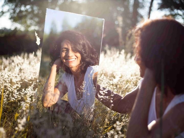 A person sits outdoors in a field of tall, pale grasses, holding a mirror that reflects their face. Warm sunlight casts a golden glow across the scene, illuminating both the person and the surrounding vegetation. The mirror shows a clear reflection of their expression, creating an introspective, serene atmosphere