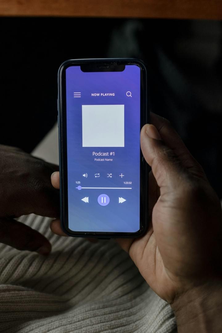 A person holds a smartphone displaying a podcast player interface. The screen shows “NOW PLAYING” at the top, followed by the title “Podcast #1” and the subtitle “Podcast name.” Standard media controls—play/pause, skip forward, and skip backward—are visible beneath the text, along with a playback progress bar. The app’s background features a smooth gradient of blue and purple hues, creating a calm and modern aesthetic. The image captures the experience of engaging with digital audio content, emphasizing themes of mobile accessibility, personal enrichment, and the integration of technology into everyday listening habits