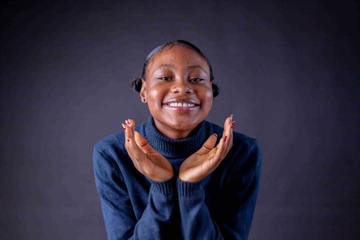 A person stands against a dark background, wearing a dark blue turtleneck sweater with their hair styled in two small buns. Their hands are raised near their face, palms upward and close together, fingers gently curved. Focused lighting highlights their face and hands, creating strong contrast with the deep background and giving the portrait a calm, expressive atmosphere.