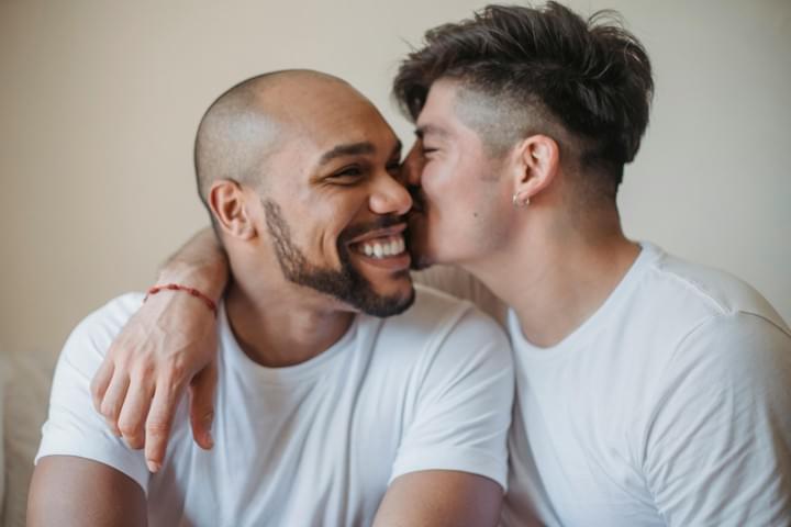 Two people sit close together indoors, both wearing white shirts. One person with short dark hair leans in to kiss the cheek of the other, who has a shaved head, a beard, and a broad smile. Their faces are close, and the moment feels warm, affectionate, and relaxed against a simple neutral background