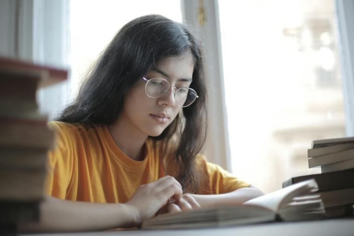 A person with long dark hair and glasses sits at a table reading a book, wearing a yellow shirt. Several books are stacked nearby, and soft natural light comes through a window behind them. The scene feels calm and focused, capturing a quiet moment of concentration and learning