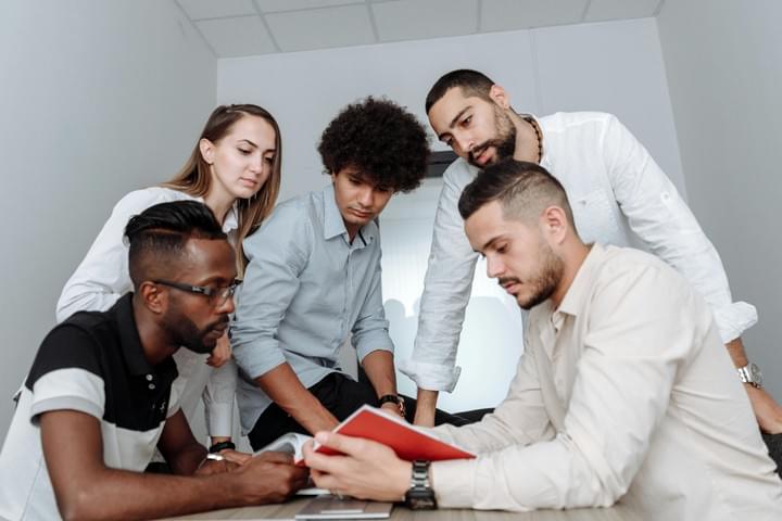 Five people stand closely together indoors, gathered around a red notebook or folder that one of them is holding. They appear to be discussing or reviewing something, leaning in with focused expressions. The setting has neutral walls and bright, even lighting, giving the scene a collaborative, team‑oriented feel.