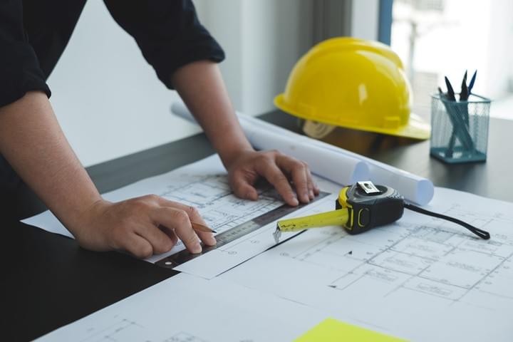 Two people are seated at a desk and are reviewing a set of building plans, the desk also has a laptop, level, and hardhat on it
