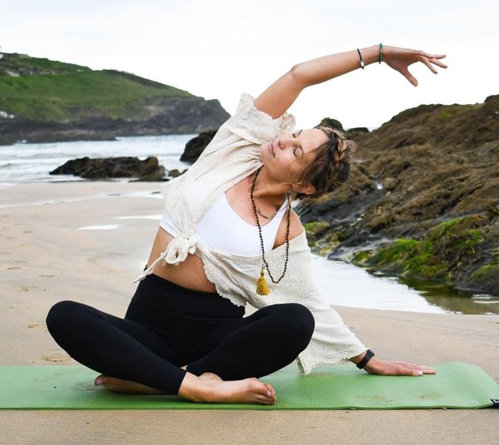 A woman is sitting crossed legged on a yoga mat, on a beach. There are green cliffs and sea rocks behind her.