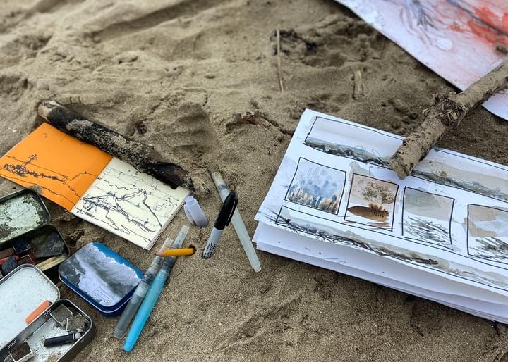 A photo showing a sketchpad with painted sketches is placed on the sand with drawing materials.