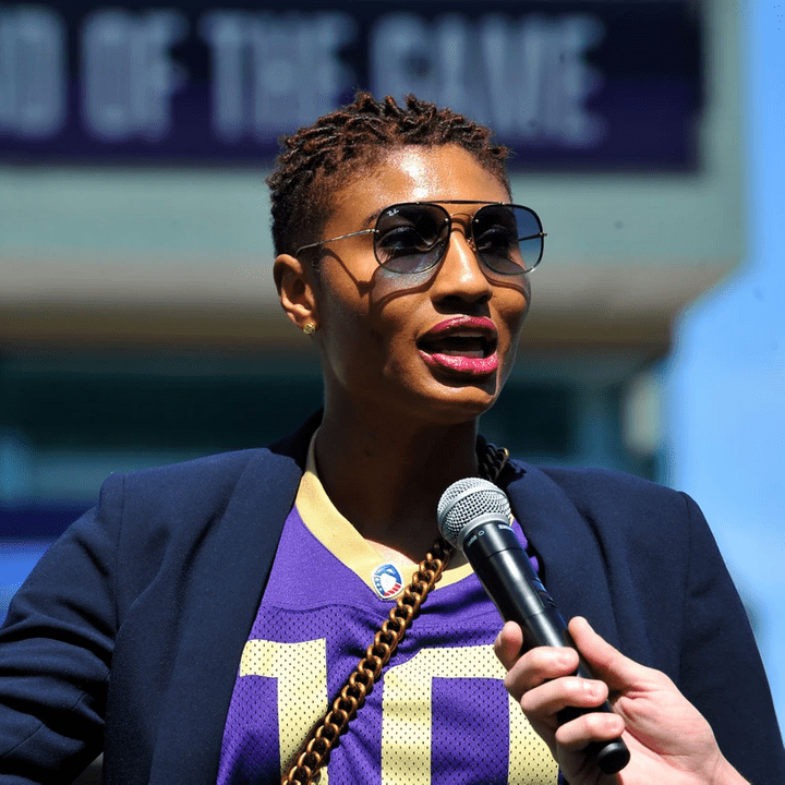 Angel speaks into microphone wearing sunglasses and magenta lipstick