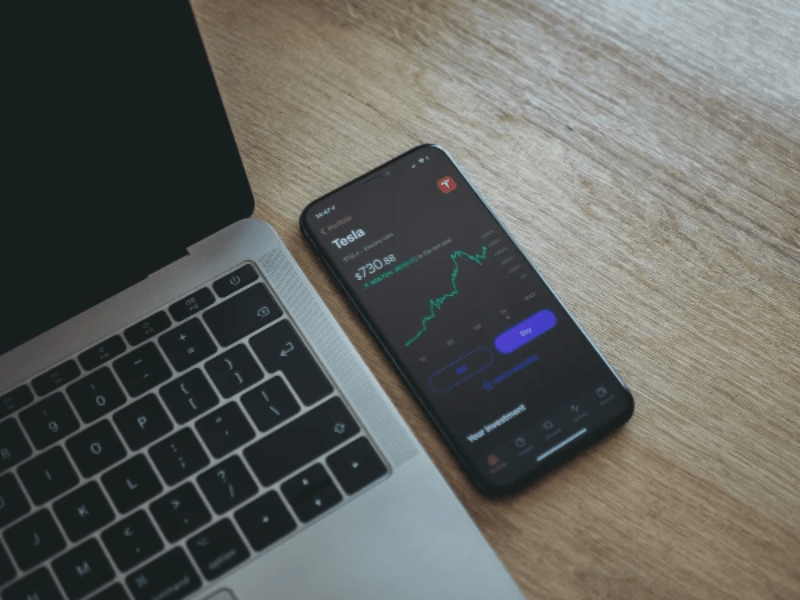 Black smartphone displaying Tesla stock chart next to a macbook laptop on a wooden table