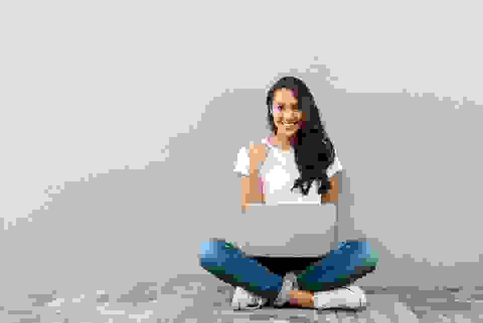 woman sat cross-legged against a wall with laptop give thumbs up