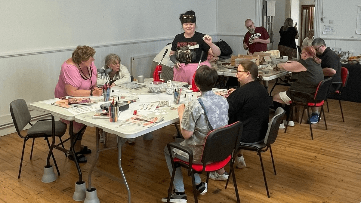 Emma is standing at the end of the table showing everyone how to use the badge making machine. She is smiling and holding a badge, whilst 4 participants sit around her and watch. They are all engrossed. The backdrop is Biggin Hall in Dover where the event was held.